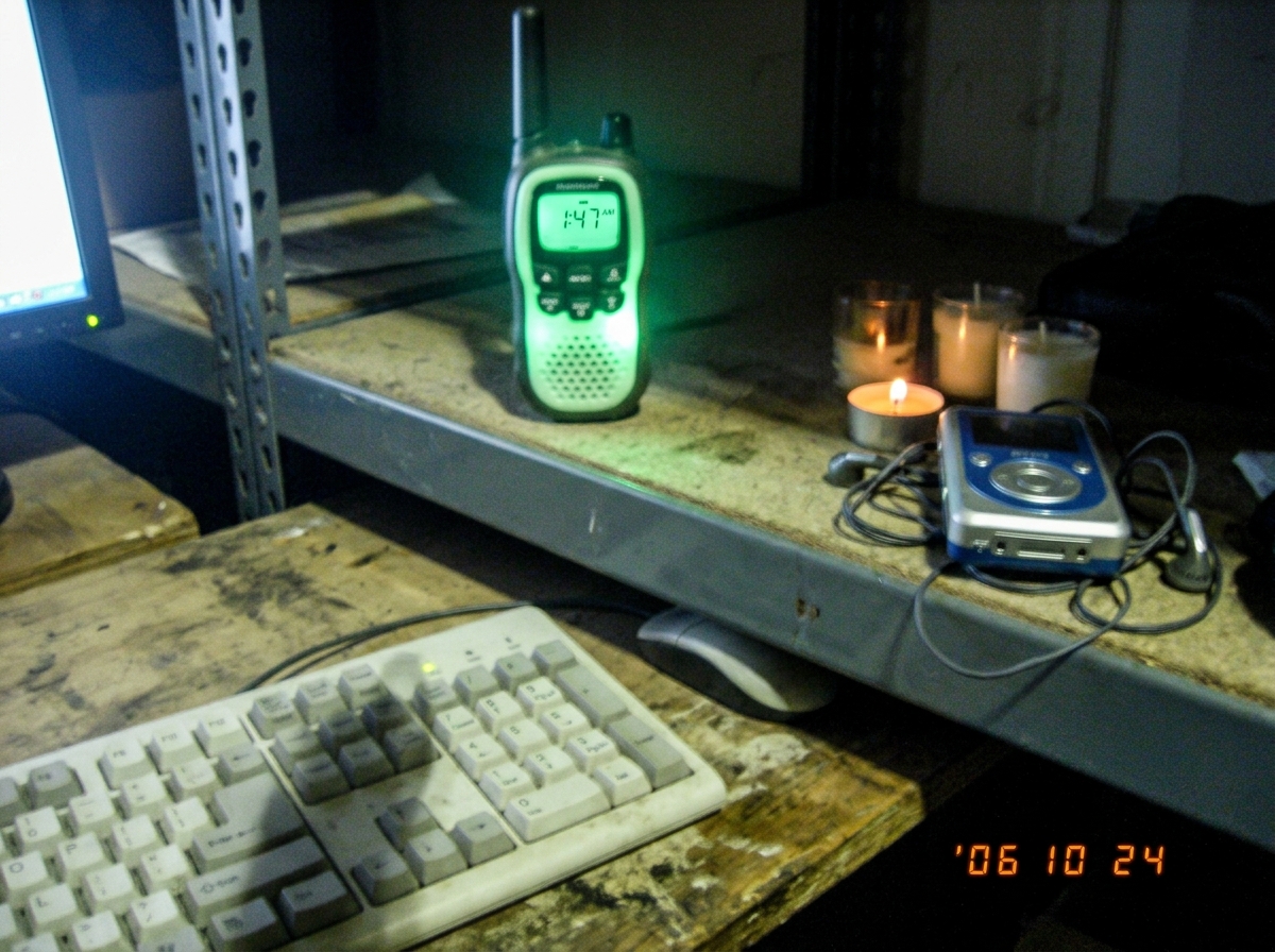 A blurry night photo of a green-glowing walkie talkie on a warehouse shelf, the digital display reading 1:47 AM, with a faint shadow of a cat on the Enter key of a keyboard in the foreground, and a small shrine of candles and an MP3 player nearby