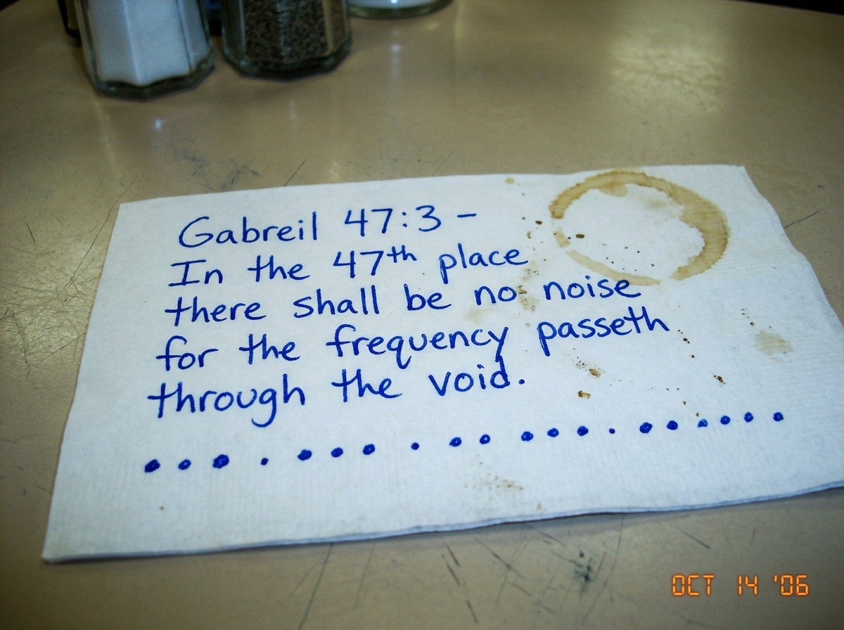 A close-up photo of a greasy truckstop diner napkin with handwritten scrawl in blue ballpoint pen. The writing reads: 'Gabreil 47:3 - In the 47th place there shall be no noise for the frequency passeth through the void.' Underneath is a diagram showing three dots, then a period, then three dots, the
