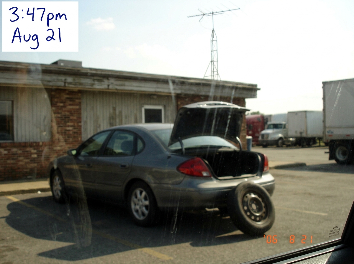 A blurry photo taken from a car window at a truckstop diner parking lot, showing a dark grey 2000s sedan with its trunk open and a spare tire leaning against the bumper. In the background a radio antenna is visible on the roof of the building. A handwritten label in the corner says '3:47pm Aug 21' i