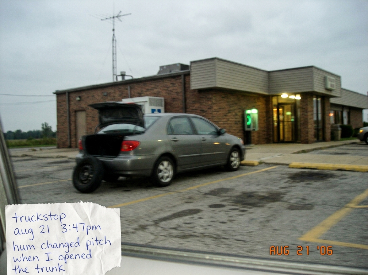 A blurry photo taken from a car window at a truckstop diner parking lot, showing a grey 2000s sedan with its trunk open and a spare tire leaning against the bumper. In the background a radio antenna is visible on the roof of the building and a payphone glows faintly green near the entrance. A handwr
