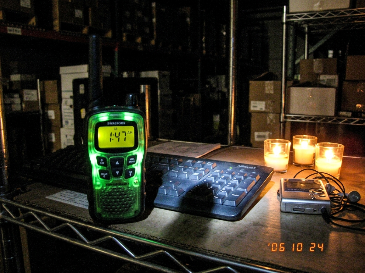 A blurry night photo of a green-glowing walkie talkie on a warehouse shelf, digital display reading 1:47 AM, faint shadow of a cat on Enter key of keyboard, small shrine of candles and MP3 player nearby