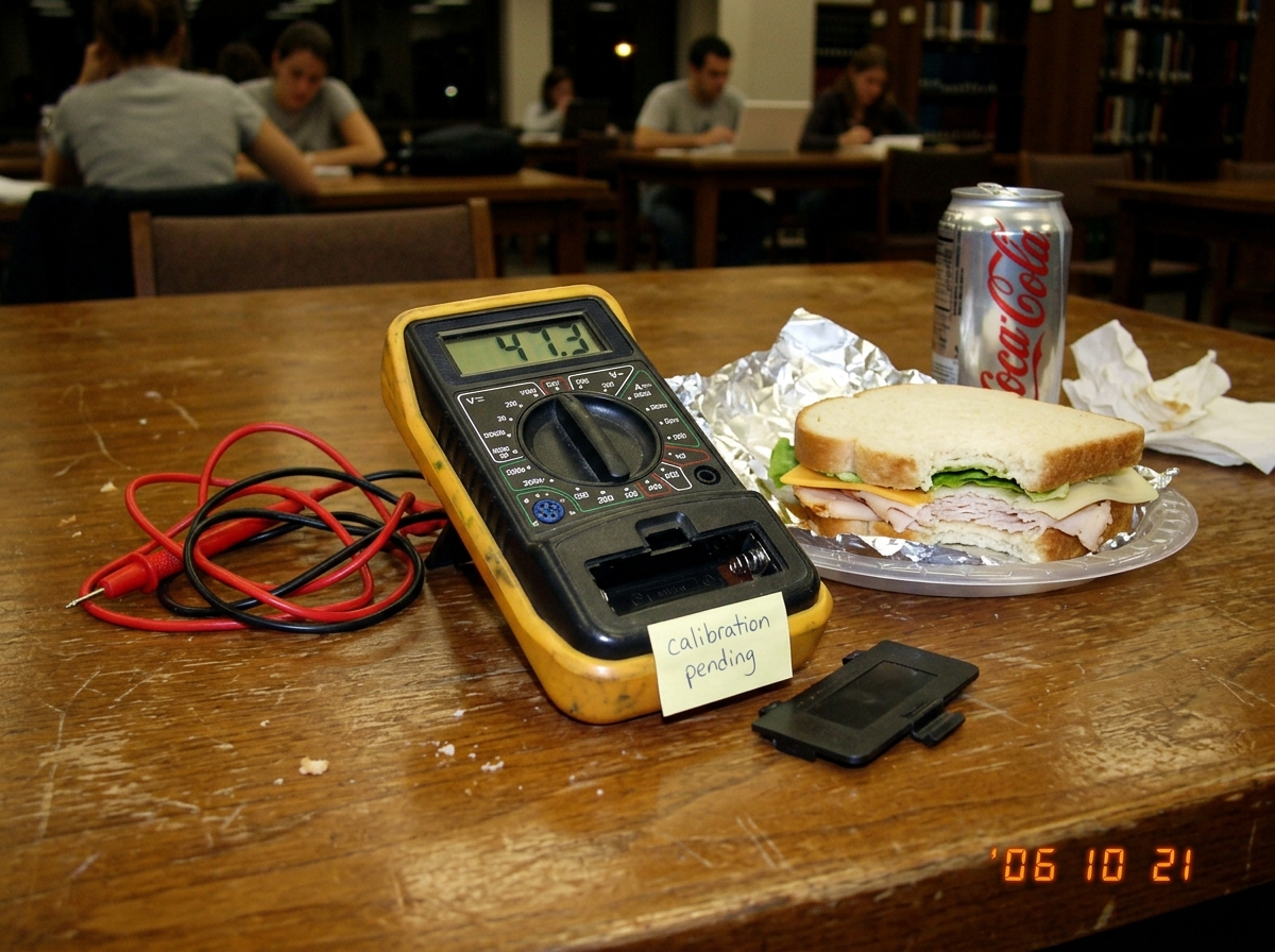 A close-up of a cheap digital multimeter lying next to a half-eaten sandwich on a library table. The multimeter display reads '47.3' but the battery compartment is open and missing a AA battery. A sticky note on the back says 'calibration pending' in messy handwriting.