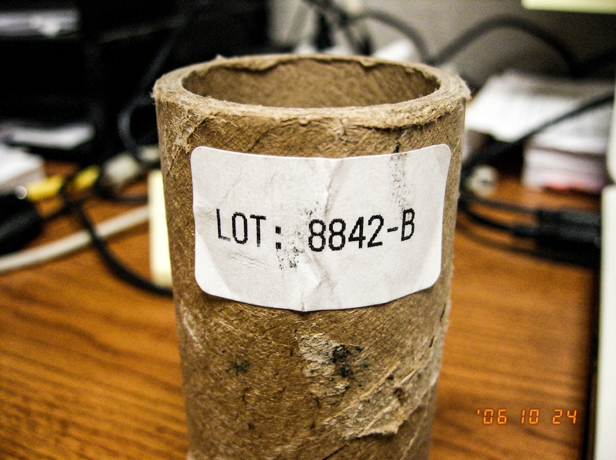 Close-up photo of a cardboard thermal paper roll tube core with a white label showing 'LOT: 8842-B' printed in black ink