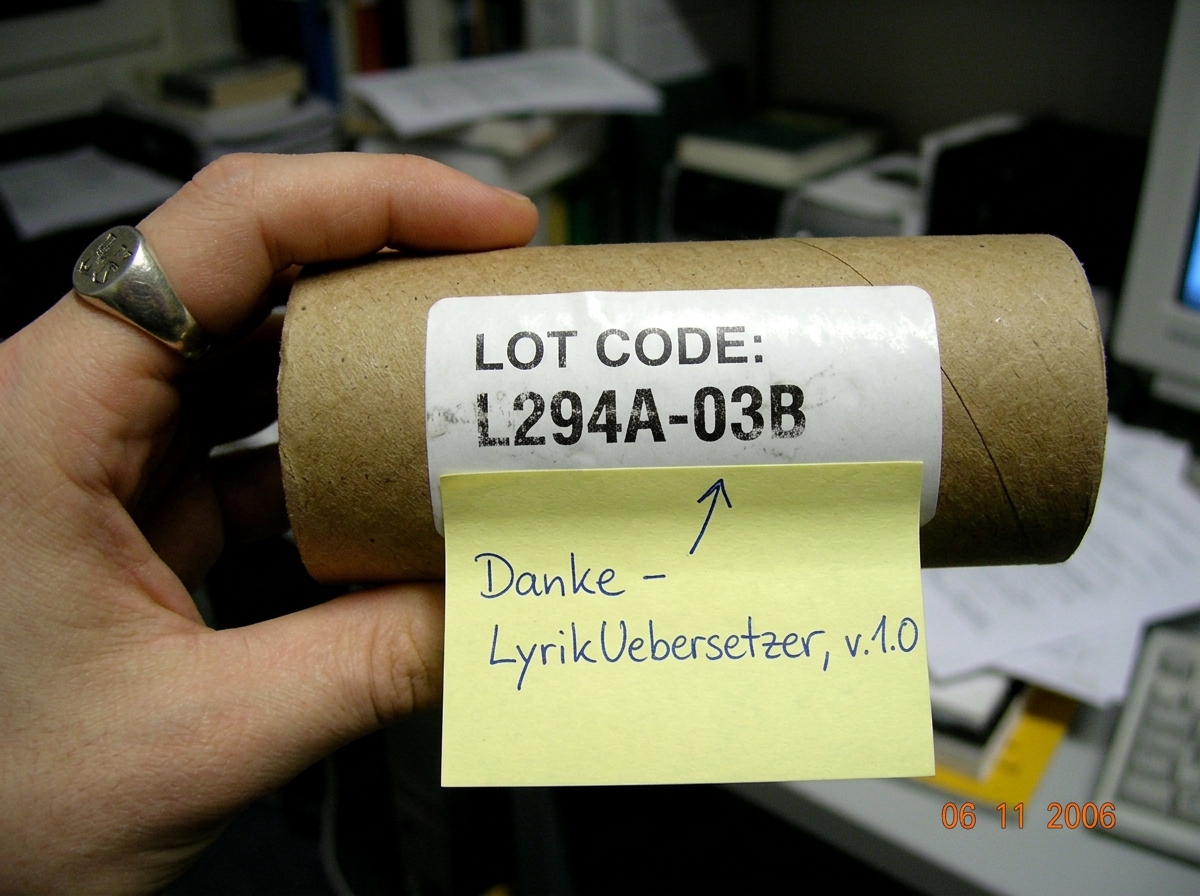 A close-up photo of a cardboard thermal paper roll tube core with a white label showing a lot code. The tube is held by a hand with a German university ring visible on the thumb. Below the label, a sticky note in neat handwriting reads 'Danke - LyrikUebersetzer, v.1.0' with an arrow pointing to the 