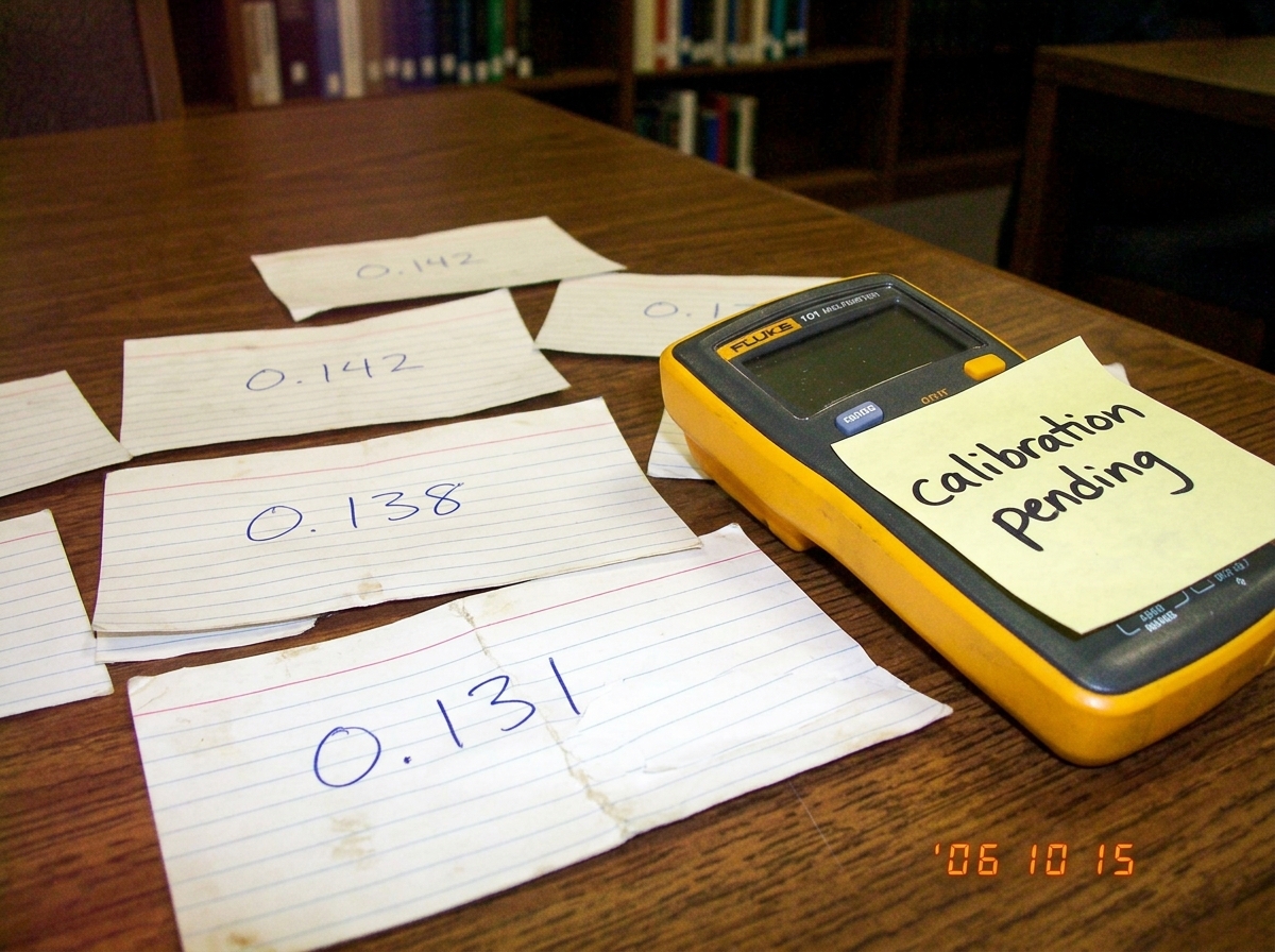A close-up photo of a library table with scattered index cards showing handwritten numbers (0.142, 0.138, 0.131) arranged next to a cheap Fluke multimeter with a dead battery. A sticky note on the multimeter reads 'calibration pending' in messy handwriting.
