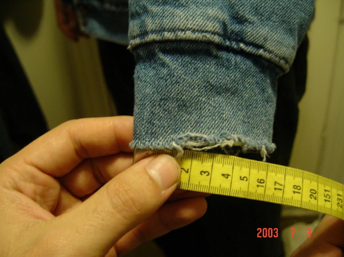 A close-up photo of a denim hoodie cuff, slightly frayed at the edge, held against a sewing measuring tape showing the thread count. The weave is clearly visible in the fabric grain. The photo has a slight blur and a date stamp in the corner reads '2003'. The lighting is dim and warm, taken indoors.