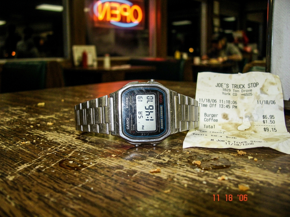 A close-up photo of a scratched Casio A158W wristwatch resting on a worn wooden truckstop diner table, the display reading 1:46, next to a crumpled thermal paper receipt. The lighting is warm and dim, with the faint reflection of a neon sign visible in the watch crystal. Dust motes float in the ambe