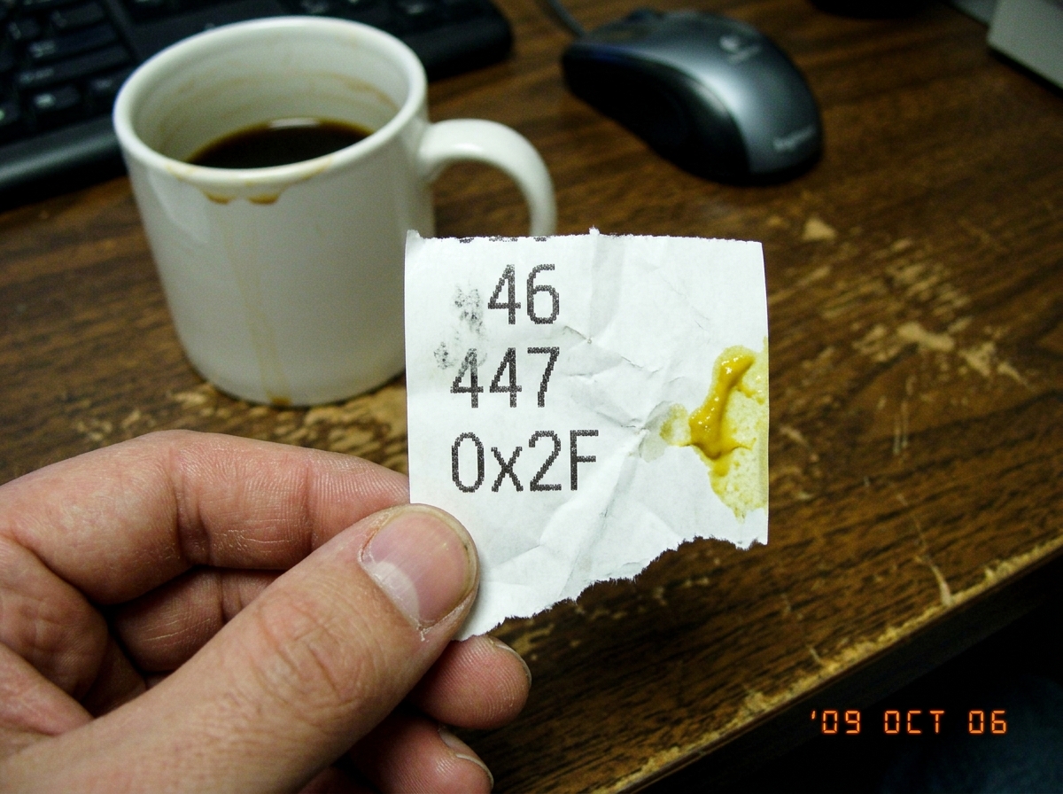A close-up photo of a thermal printer receipt, torn and slightly crumpled, showing three numbers printed in sequence: '46', '47', and '0x2F' in hexadecimal. Next to the receipt is a yellow mustard stain on the paper at the edge, positioned near where byte 0x2E would be. The receipt is held in someon