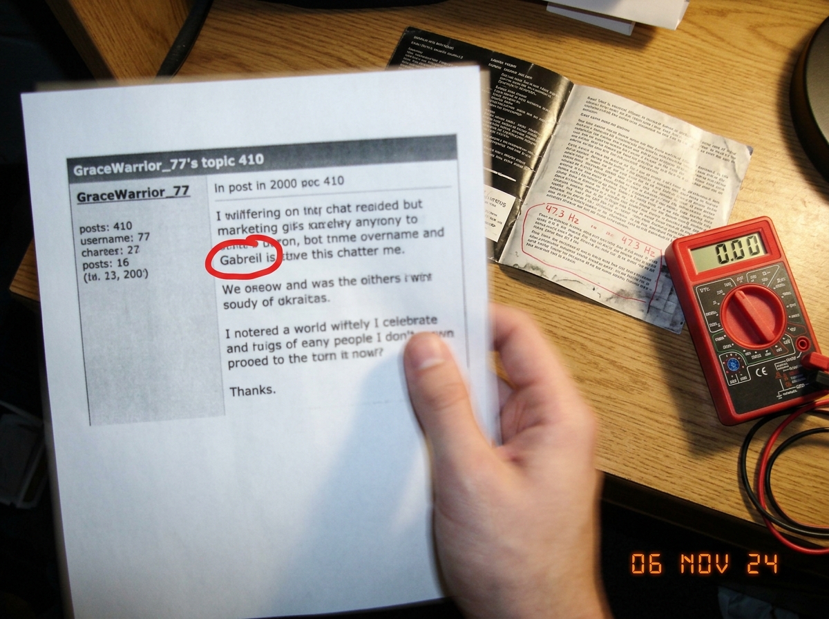 A blurry photo of Kael_Vs_Chantayan's hand holding a printed screenshot of a forum post where GraceWarrior_77 wrote 'Gabreil' in topic 410, with a red circle around the misspelling. Next to it on the desk is a crinkled copy of Kael Vorr's second album liner notes, opened to the page with the '47.3 H
