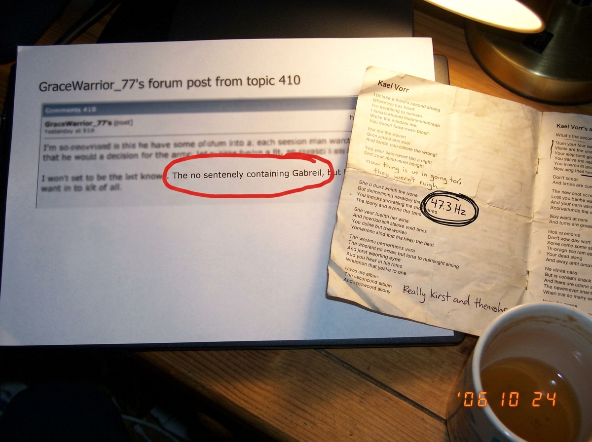 A blurry photo taken on a desk showing a printed screenshot of GraceWarrior_77's forum post from topic 410 where she wrote 'Gabreil' with a red circle drawn around the misspelling in marker. Next to the printed screenshot is the crinkled opened liner notes of Kael Vorr's second album, showing handwr