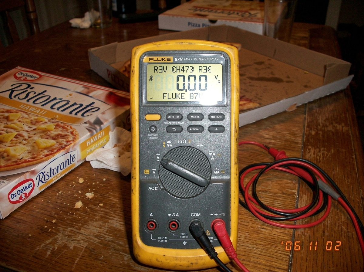 A blurry photo taken on a flip phone showing a Fluke 87V multimeter screen with garbled, half-formed characters on the firmware version line. The meter is sitting on a table with a pizza box visible in the background. There's a frozen pizza box (pineapple) partially in frame.