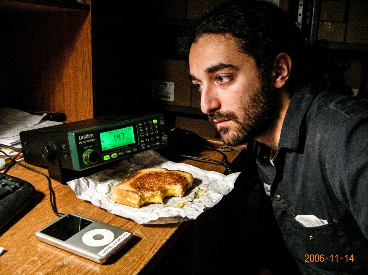 A green-glowing police scanner sitting on a warehouse desk at night, its screen showing '1:47' in glowing digits, next to a half-eaten cheese sandwich and an old MP3 player with Gabriel's name sticker.