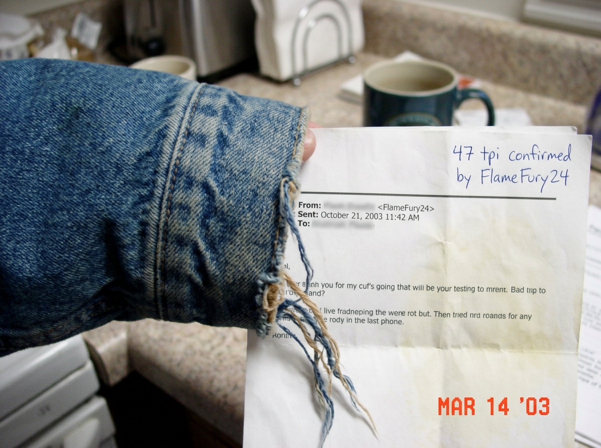 A blurry photo of a denim jacket cuff with loose threads visible, held up next to a printed email from 2003. A handwritten note in the corner says '47 tpi confirmed by FlameFury24'. The photo is taken on a kitchen counter with a coffee mug in the background.