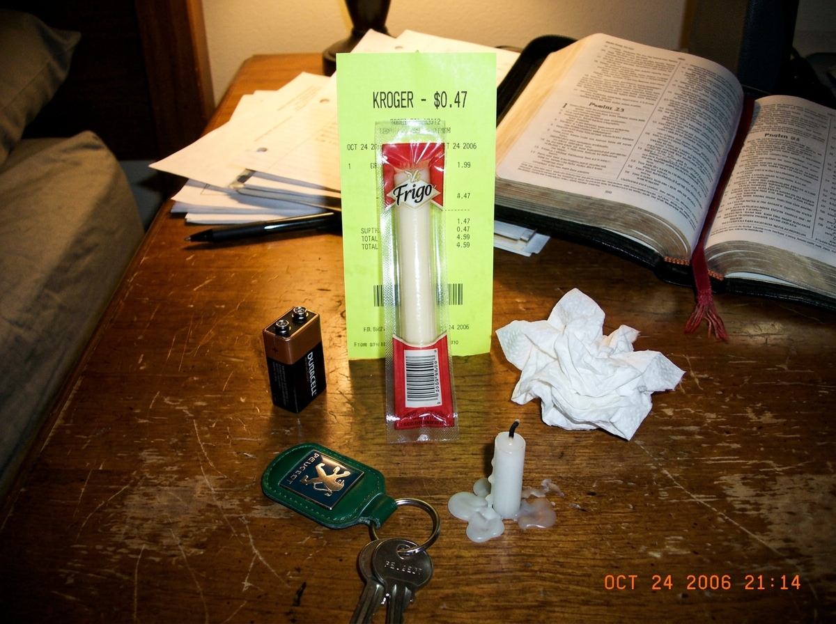 A photograph taken in dim evening lamplight: a small homemade shrine on a wooden desk. The shrine consists of a single string cheese stick, still in its plastic wrapper, propped upright against a bright fluorescent receipt that reads 'KROGER - 0.47' at the top. The cheese is surrounded by a circle o