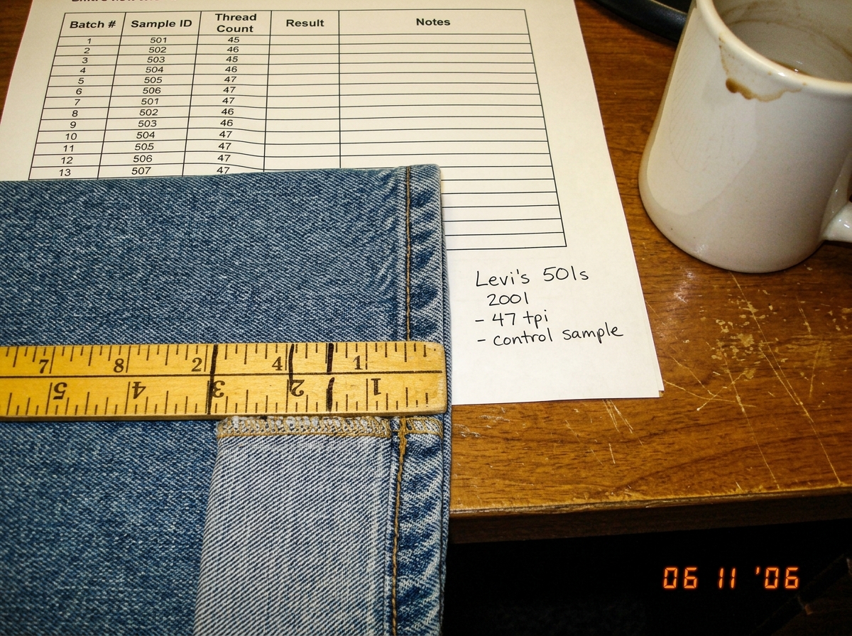 A close-up photo of a denim jean cuff laid flat on a desk with a yellow fabric ruler across the weave, showing the thread count aligned to 47 threads per inch. Next to the ruler is a printed spreadsheet with column headers visible and a handwritten note saying 'Levi's 501s 2001 - 47 tpi - control sa