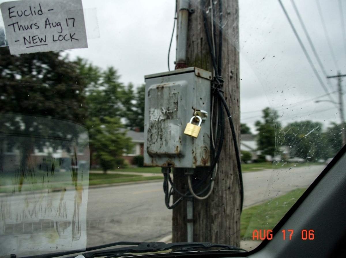 A blurry photo taken from a car window showing a gray electrical junction box on a utility pole, with a shiny new brass padlock visible on the latch. A handwritten note in the corner says 'Euclid - Thurs Aug 17 - NEW LOCK'. The photo is slightly out of focus and shows the reflection of a windshield.
