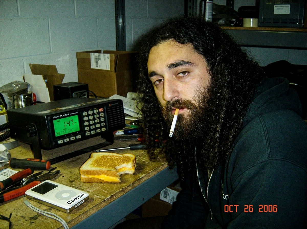 A green-glowing police scanner on a warehouse desk at night, showing 1:47 on its screen, with a half-eaten cheese sandwich and MP3 player with Gabriel sticker