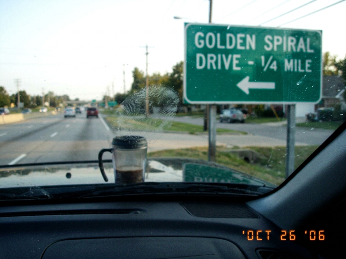 A blurry flip-phone photo taken through a windshield of a road sign on the way home from Kroger. The sign reads 'GOLDEN SPIRAL DRIVE - 1/4 MILE' with an arrow, but the arrow is pointing the wrong way - back toward the highway instead of into the neighborhood. Taken on a Motorola RAZR, so the photo i