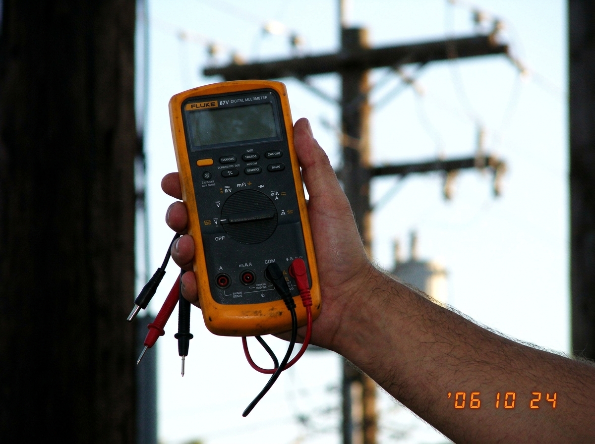 A hand holding a Fluke multimeter with probes dangling uselessly against a blurred background of a utility pole