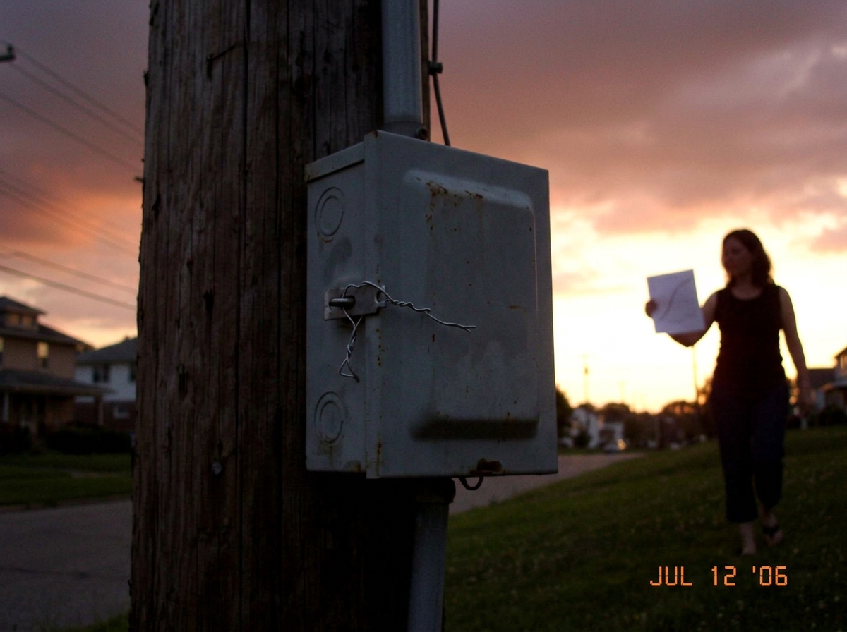 A photo from July 12, 2006 at dusk near a utility pole in Euclid Ohio. A gray electrical junction box on the pole has a twisted wire tie holding the latch closed instead of a padlock. In the background, a woman's silhouette is visible holding a piece of paper (a gamma curve chart) with the evening s