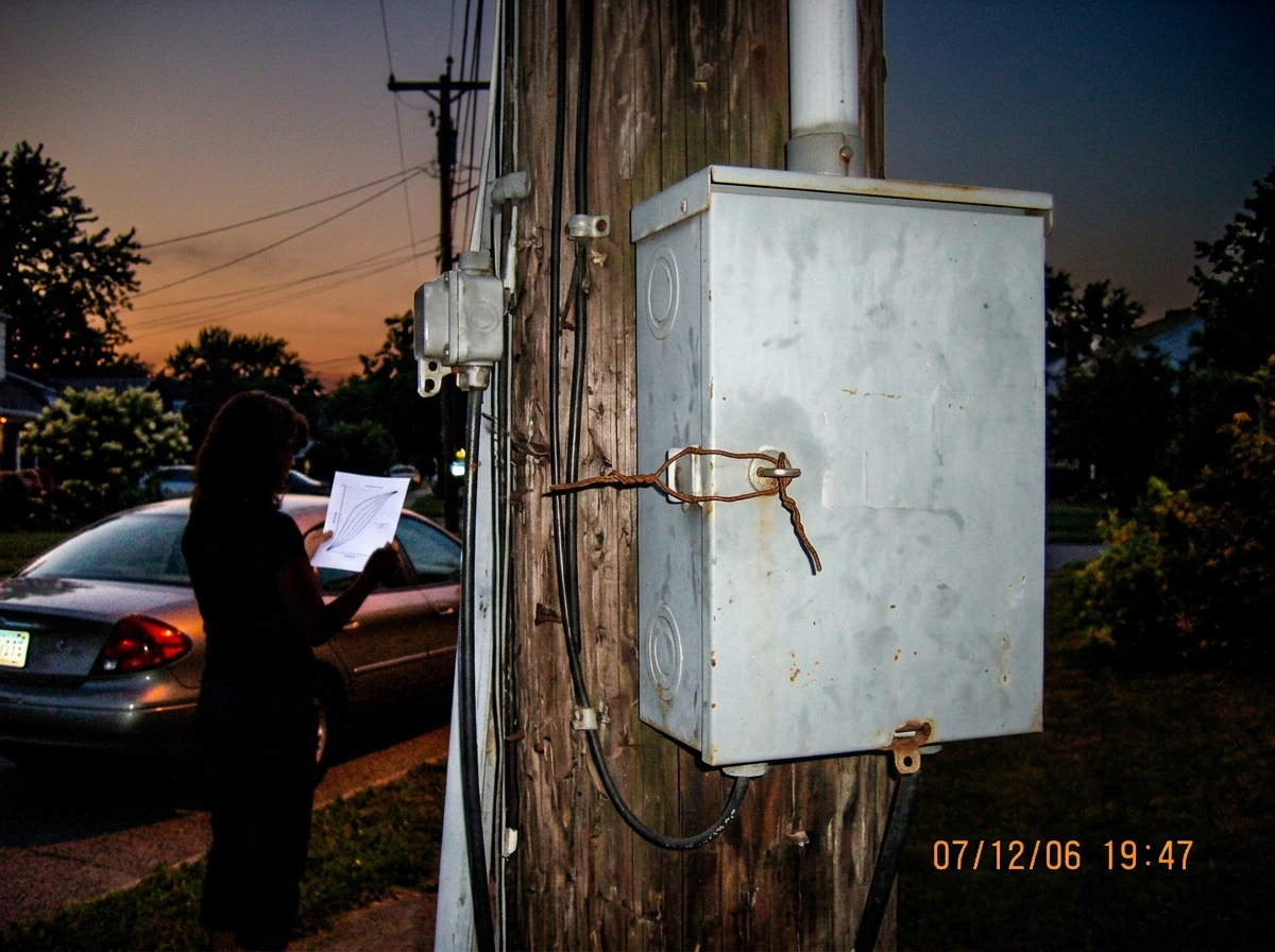 A photo from July 12, 2006 at dusk near a utility pole in Euclid Ohio. A gray electrical junction box on the pole has a twisted wire tie holding the latch closed instead of a padlock. In the background, a woman's silhouette is visible holding a piece of paper (a gamma curve chart) with the evening s
