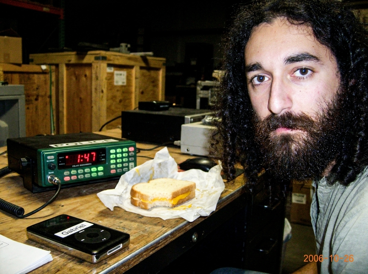 A green-glowing police scanner on a warehouse desk at night, showing 1:47 on its screen, with a half-eaten cheese sandwich and MP3 player with Gabriel sticker