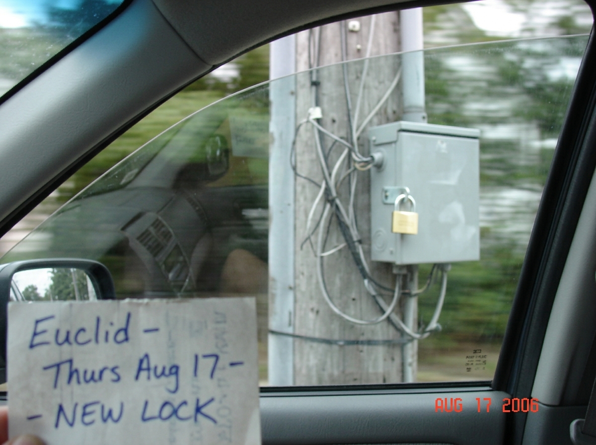 A blurry photo taken from a car window showing a gray electrical junction box on a utility pole, with a shiny new brass padlock visible on the latch. A handwritten note in the corner says 'Euclid - Thurs Aug 17 - NEW LOCK'. The photo is slightly out of focus and shows the reflection of a windshield.