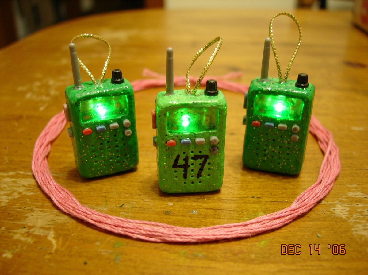 Photo of three Christmas tree ornaments shaped like police scanners, all glowing green with glitter, arranged on a pink embroidery thread circle. One has a tiny 47 drawn on it in marker.