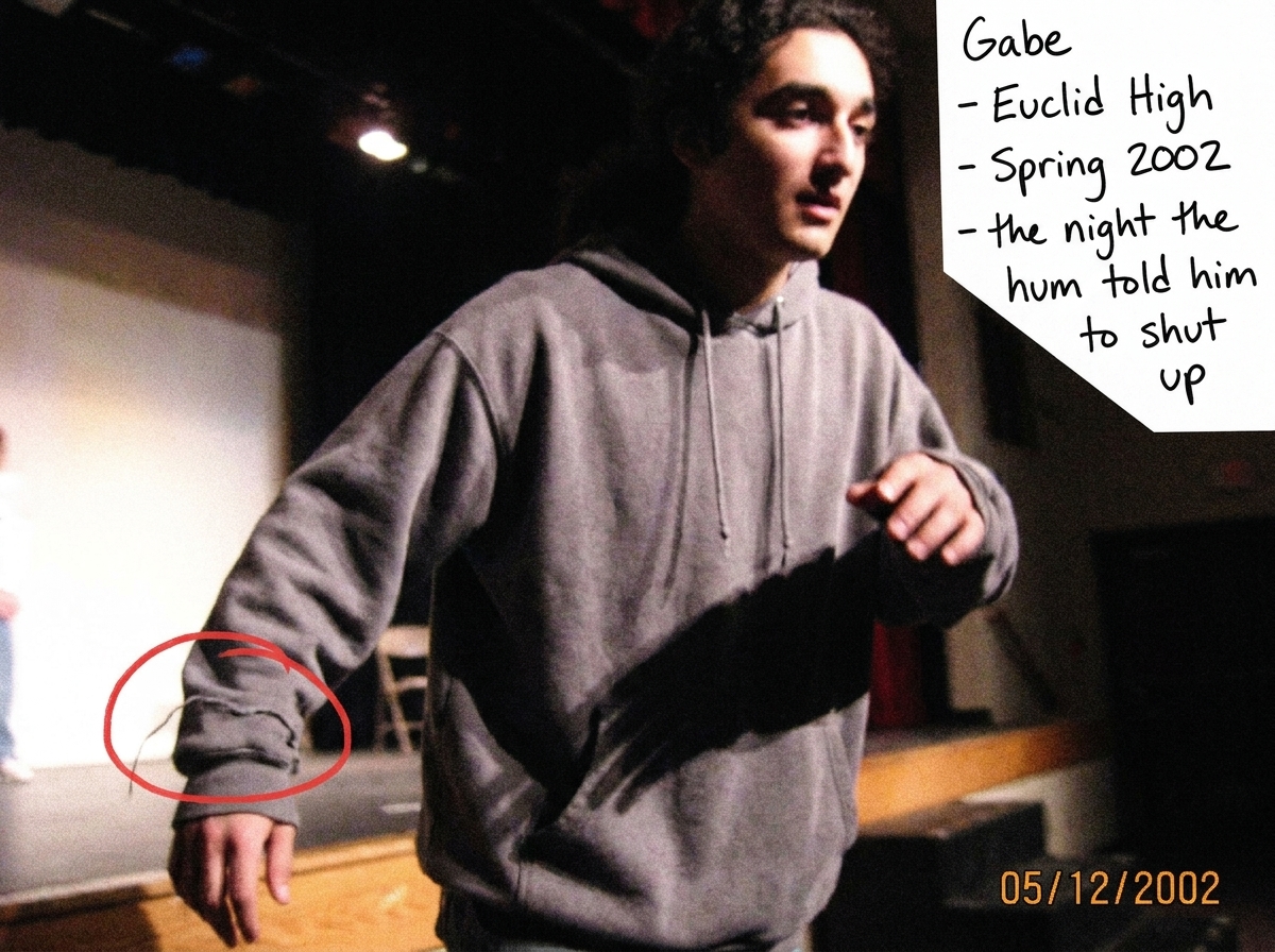A blurry high school talent show photo from 2002. A teenage boy (Gabriel Chantayan, about 16-17) is caught mid-stumble on stage, wearing an oversized gray hoodie. A loose thread from his left cuff is visible in the spotlight, circled in red marker. The stage is dark with amateur lighting. Handwritte