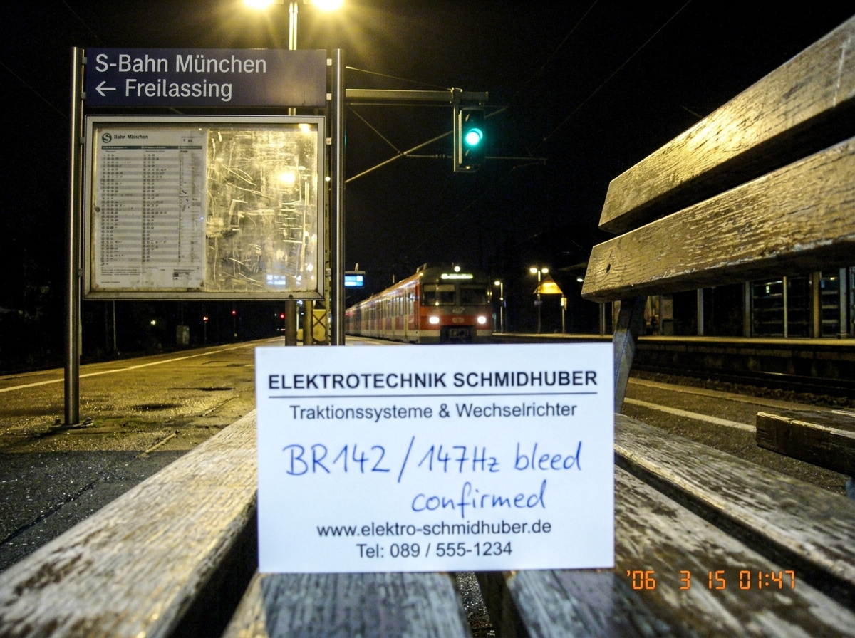A photograph taken on a train platform at night in a German suburb. A tram/S-Bahn sign reads 'S-Bahn München - Freilassing' with a schedule behind scratched plexiglass. A green-glowing overhead wire junction box is visible in the background. In the foreground on the bench is a business card for 'Ele