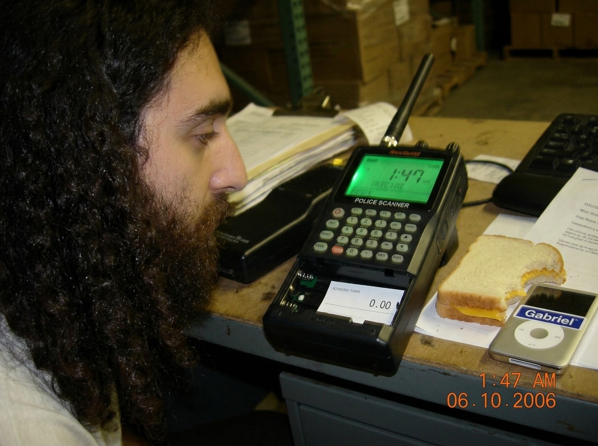 A green-glowing police scanner on a warehouse desk at night, showing 1:47 on its screen, with a half-eaten cheese sandwich and MP3 player with Gabriel sticker. The glow is visible even though there are no batteries in the compartment, which is shown open and empty with a small slip of paper that rea