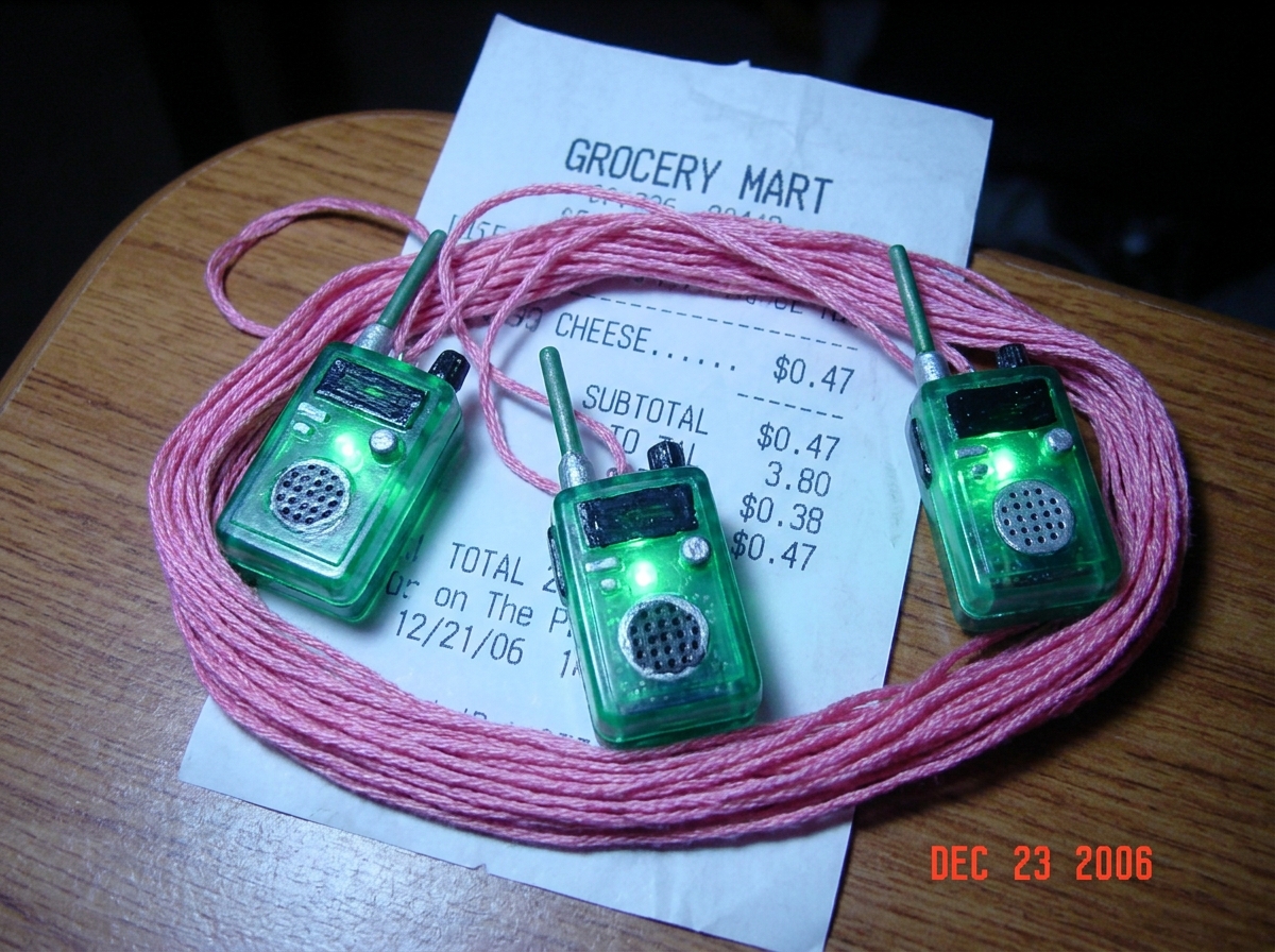Photo of three green-glowing Christmas tree ornaments shaped like police scanners, arranged on pink embroidery thread with a cheese receipt showing 47 cents