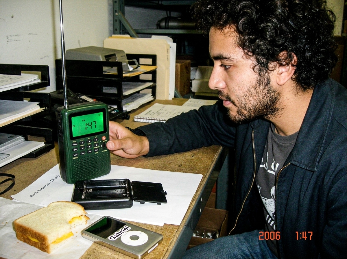 A green-glowing police scanner on a warehouse desk at night, showing 1:47 on its screen in FM mode. The FM indicator light is visible, the battery compartment is open and empty, and a small slip of paper reads 'abuela frequency - FM only'. A half-eaten cheese sandwich and MP3 player with Gabriel sti