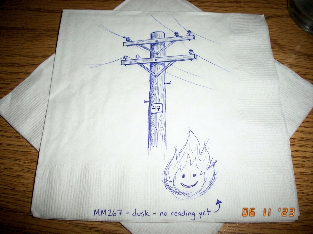 A ballpoint pen drawing on a white paper napkin with faint blue ink. The drawing shows a wooden utility pole with crossbars, and the number '47' is clearly written on a small rectangular tag attached to the pole. Beneath the pole, someone has scribbled a small cartoon flame with a smiley face inside