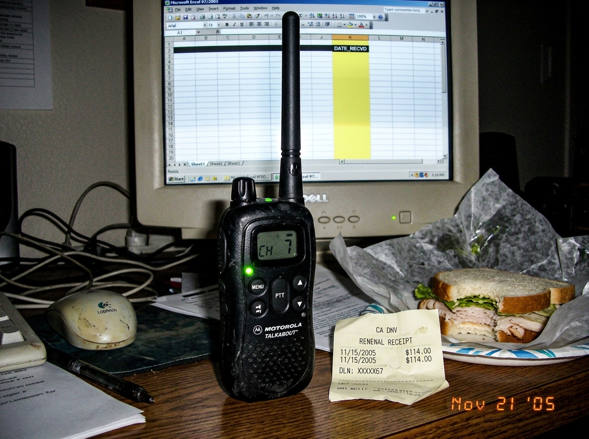 A grainy, low-res digital photo from a point-and-shoot camera circa 2005. It shows a cheap, black rubberized walkie-talkie sitting on a cluttered desk next to a half-eaten turkey sandwich and a crumpled DMV receipt. The walkie has a small green LED lit up on the front. In the background, slightly ou