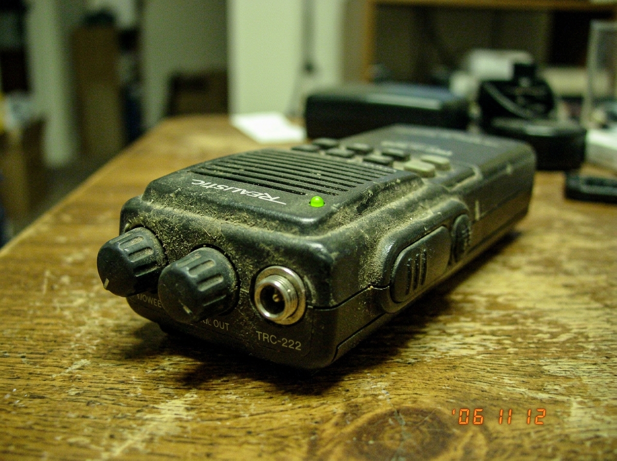 A close-up of a vintage walkie-talkie's front panel with a single green LED glowing faintly, placed on a wooden table with a faint greenish tint to the lighting. The walkie is slightly dusty, suggesting it has been handled often. The background is blurred to keep focus on the device.
