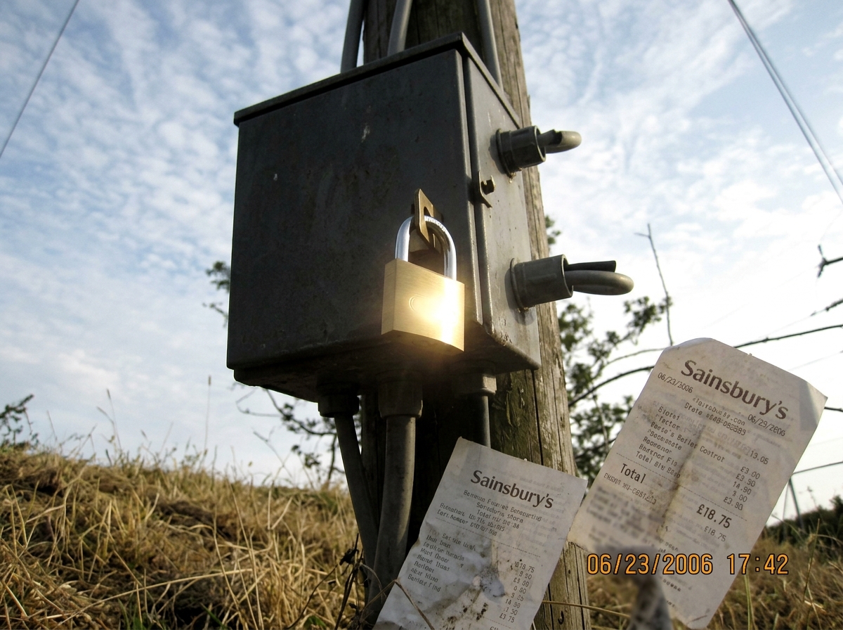 The photo is taken from about six feet away at a slight upward angle. It shows a gray electrical junction box mounted on a utility pole against a late-afternoon sky with thin clouds. A new brass padlock hangs from the hasp on the box door. The lock body catches the sun at an angle, creating a strong