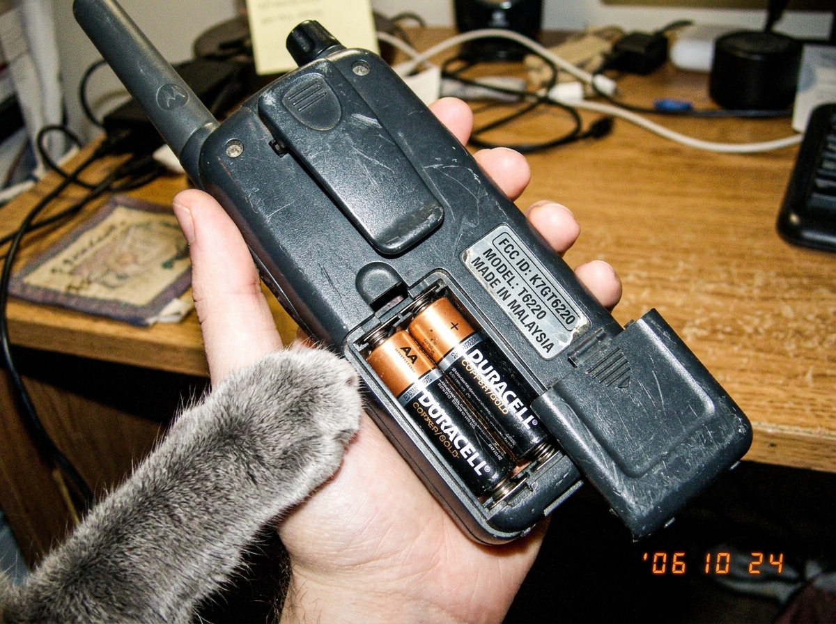 A close-up photograph of a vintage walkie-talkie held at an angle showing the back panel. The battery compartment cover is visible and partially open, revealing two AA batteries inside. The FCC ID sticker is legible on the side panel. A gray cat's paw reaches into the frame from the bottom left corn