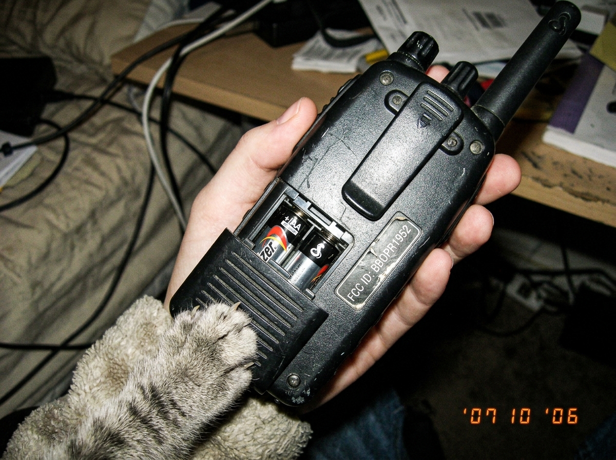 A close-up photograph of a vintage walkie-talkie held at an angle showing the back panel. The battery compartment cover is visible and partially open, revealing two AA batteries inside. The FCC ID sticker is legible on the side panel. A gray cat's paw reaches into the frame from the bottom left corn