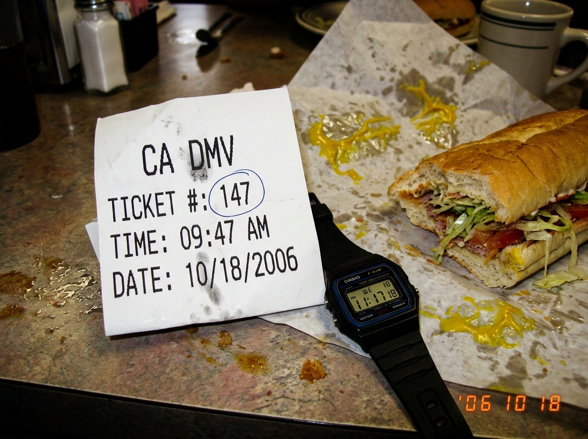 A crumpled DMV ticket receipt on a greasy diner table. The number is 147, timestamp 9:47 AM. Next to it sits a half-eaten footlong sub sandwich with mustard smeared aggressively across the wrapper. A Casio digital watch shows 11:17.