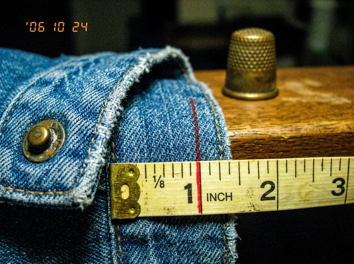 A close-up macro photograph of a blue denim jacket cuff, taken under harsh fluorescent lighting. The weave of the denim is clearly visible, with individual cotton threads crossing at right angles. A sewing tape measure with inch markings lies beside the fabric, and a red line has been drawn along a