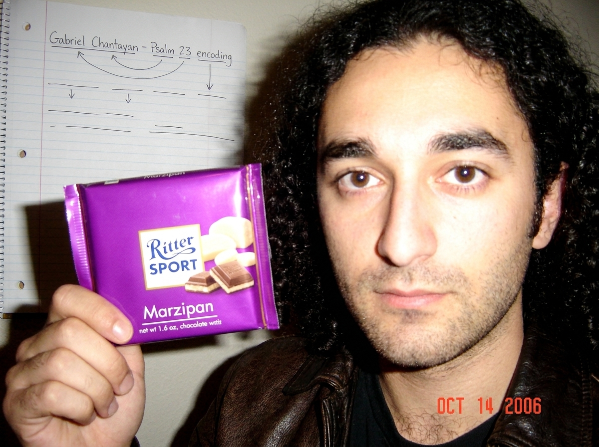 A Ritter Sport chocolate wrapper, purple with the words 'Marzipan' on it, being held up against a blurry background of a dorm room wall with a notebook page visible that has 'Gabriel Chantayan - Psalm 23 encoding' scrawled in handwriting with arrows and underlines.