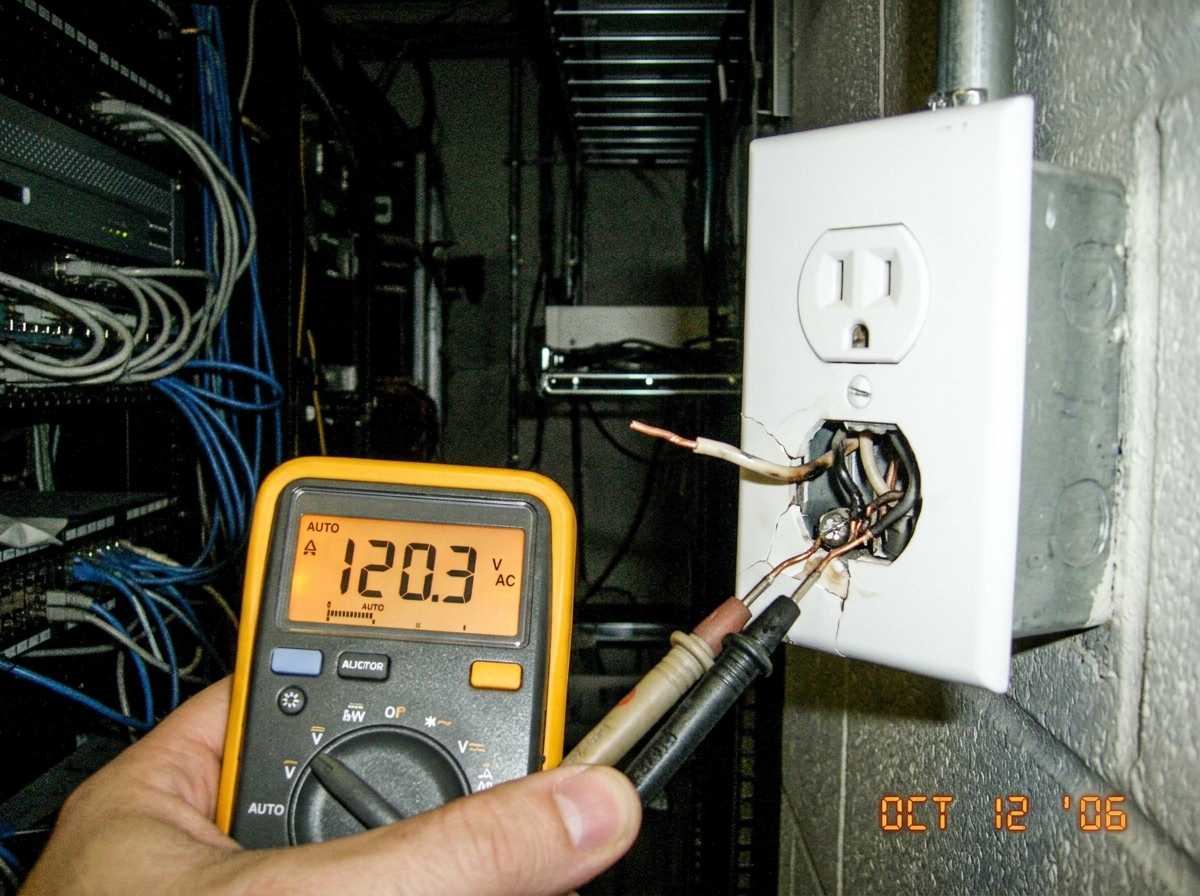 A close-up of a cracked wall socket with exposed wiring and a multimeter reading 120V AC, taken in a dimly lit server room