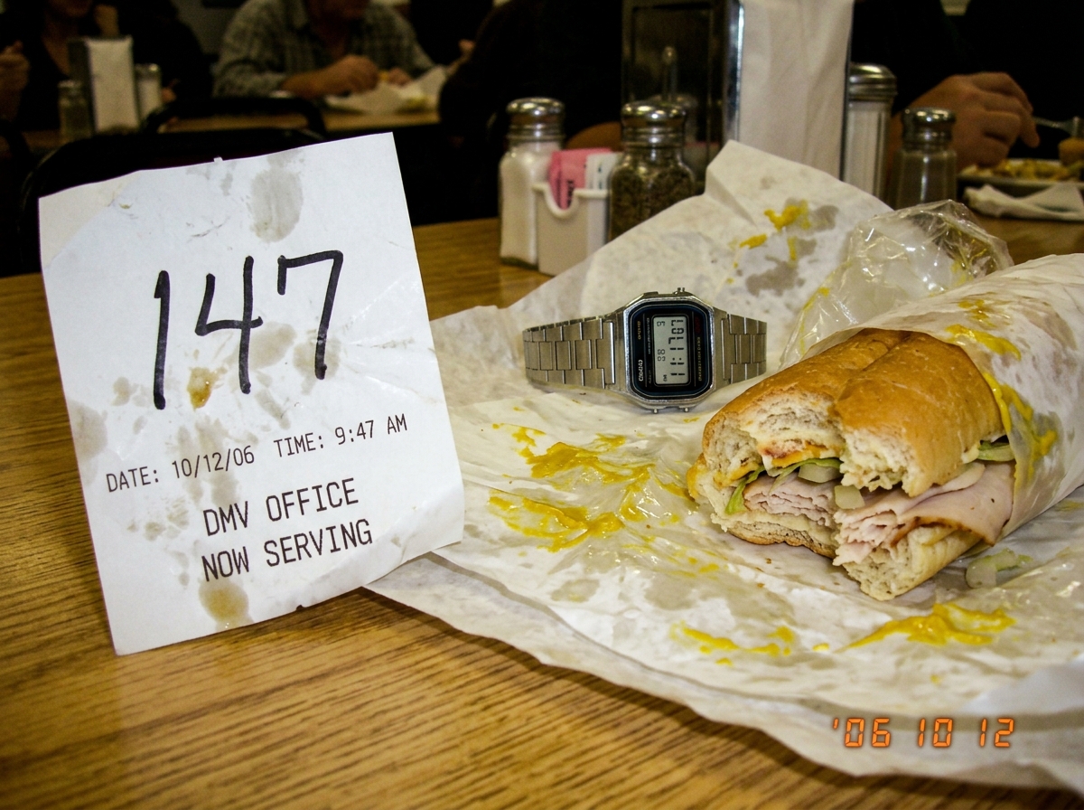 A crumpled DMV ticket receipt on a greasy diner table showing number 147 with timestamp 9:47 AM. Next to it sits a half-eaten footlong sub sandwich with mustard smeared aggressively across the wrapper. A silver Casio digital watch shows 11:17. The photo is slightly blurry and clearly taken in a hurr