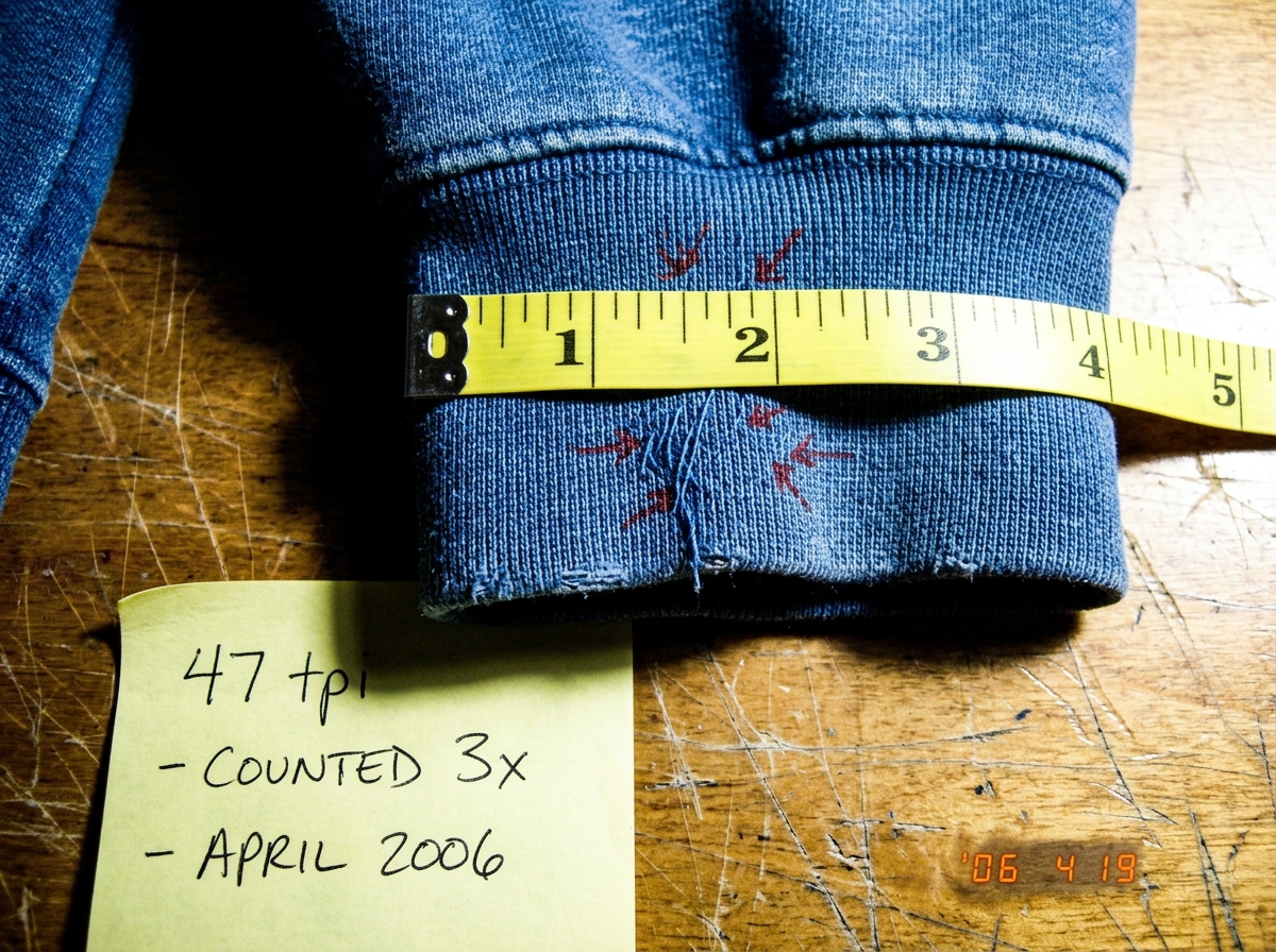 A close-up macro photograph of a denim hoodie cuff, taken on a wooden sewing table. A yellow sewing tape measure lies across the fabric showing the inch markings. Small red arrows drawn in marker point to individual blue cotton threads crossing vertically and horizontally. A handwritten sticky note