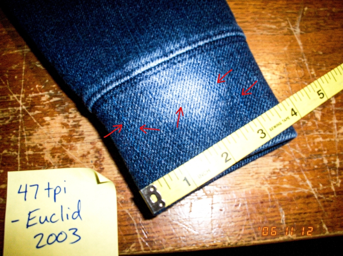 A close-up macro photo of a denim hoodie cuff laid flat on a wooden sewing table. A yellow sewing tape measure crosses diagonally over the fabric showing inch markings. Small red hand-drawn arrows point to individual blue threads. A handwritten sticky note in the corner reads '47 tpi - Euclid 2003' 