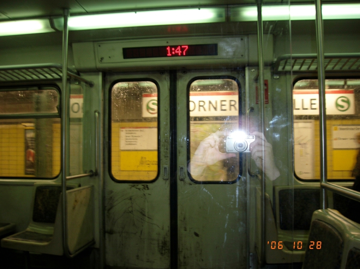 A blurry photo taken from inside a Berlin S-Bahn train at night. The digital display above the doors shows '1:47' in white LED digits. Through the window, a platform sign is visible showing the station name partially obscured, with German text. The reflection of the photographer's hand holding a dis