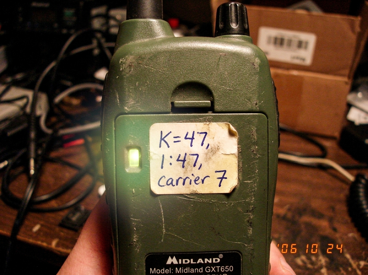Close-up photo of the same green-glow tactical walkie talkie showing a small sticker on the back with handwritten numbers 'K=47, 1:47, carrier 7', the green LED casting a faint glow on the paper sticker