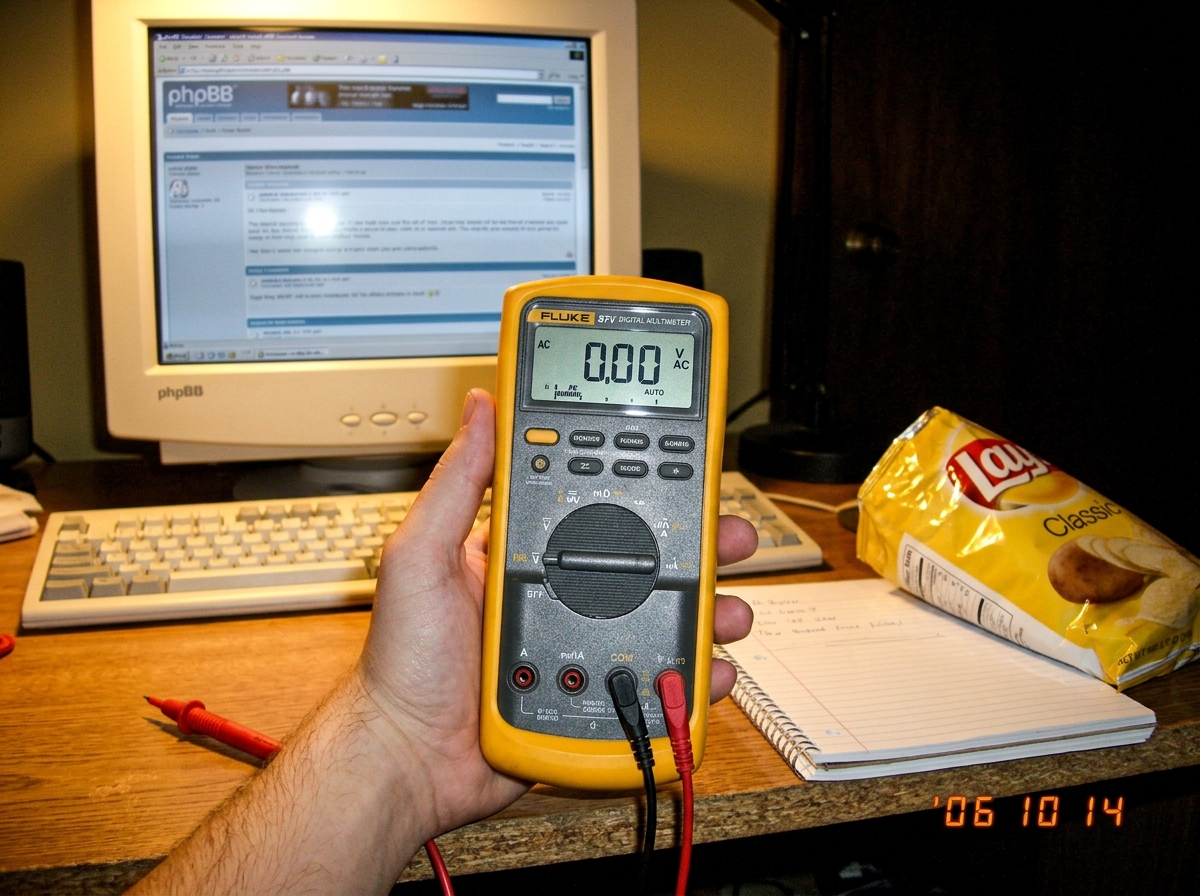 A close-up photo of a Fluke 87V multimeter held in one hand, with the test leads dangling, resting on a wooden desktop next to a half-eaten bag of chips and a spiral notebook. The meter is turned on and set to AC voltage, reading 0.00. In the background, a CRT monitor shows a phpBB forum page. The l