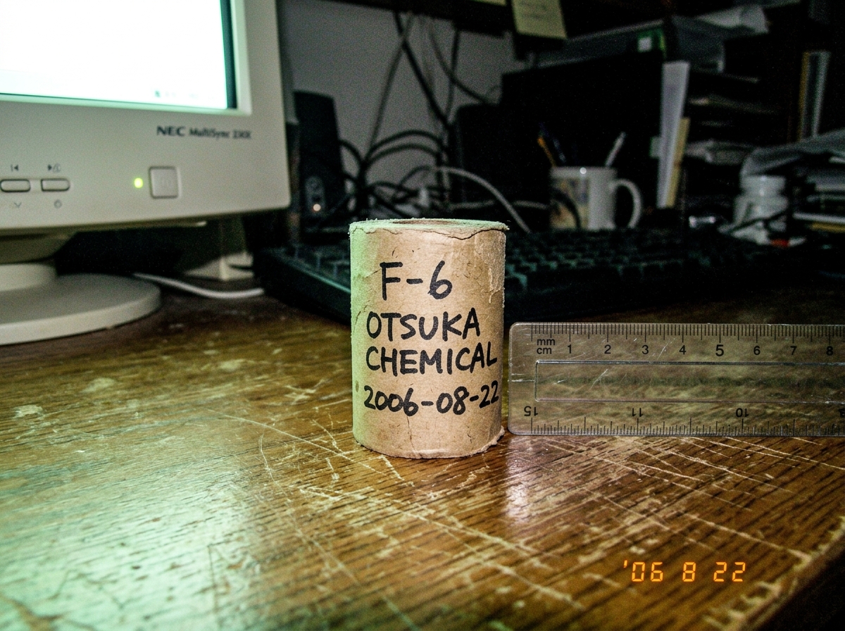 A close-up shot of a thermal paper roll core labeled 'F-6 Otsuka Chemical 2006-08-22' sitting on a wooden desk next to a ruler for scale, with a faint green glow from a nearby CRT monitor casting a subtle hue on the paper.